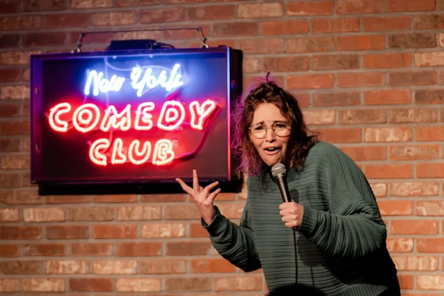 Beth on stage with a mic under a red neon New York Comedy Club sign
