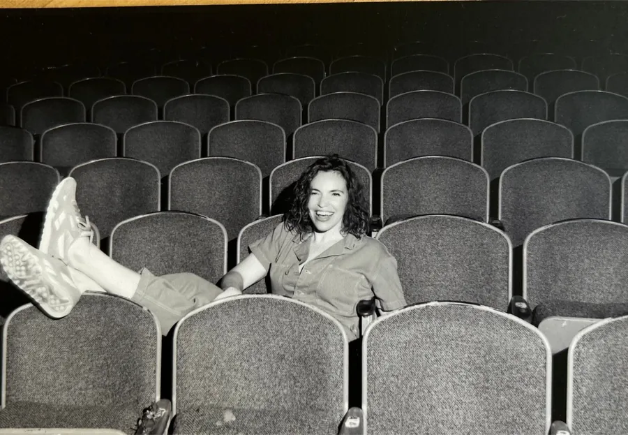 Postcard, design 1: Beth laughing with her feet up across theater seats, black and white