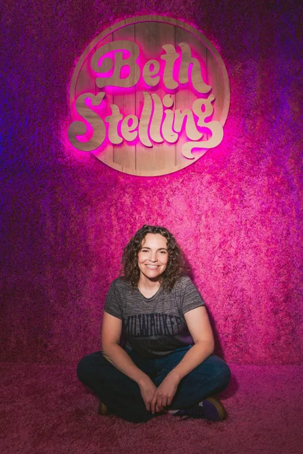 Beth sitting cross-legged under a pink neon sign of her name