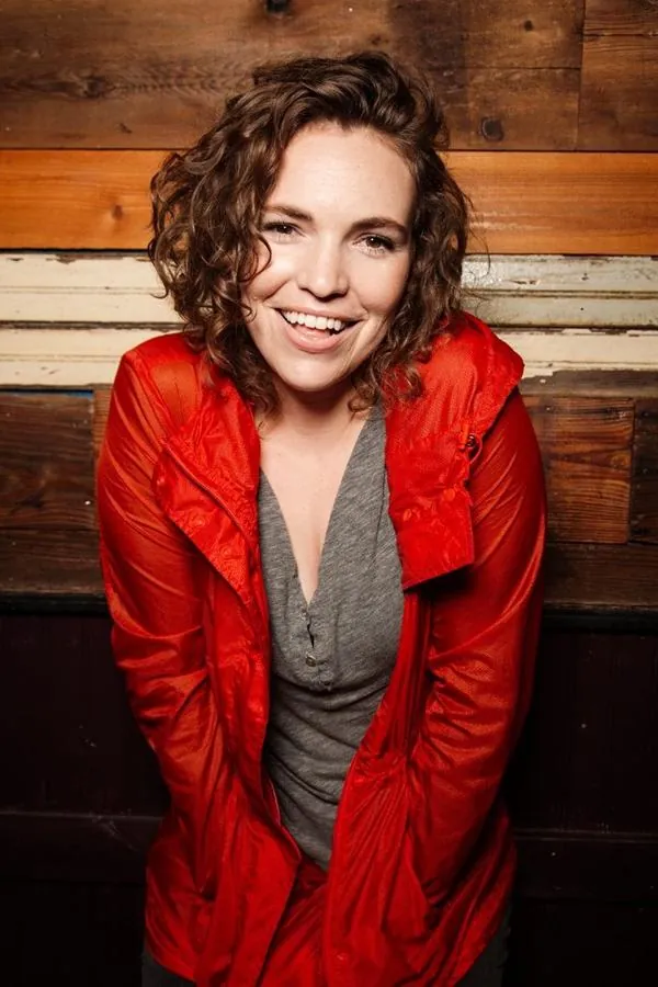Beth in a red coat smiling against a wooden wall