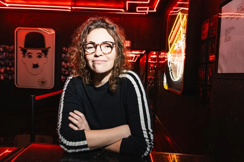 Beth in a black track top smiling in a red-lit room at the Comedy Store
